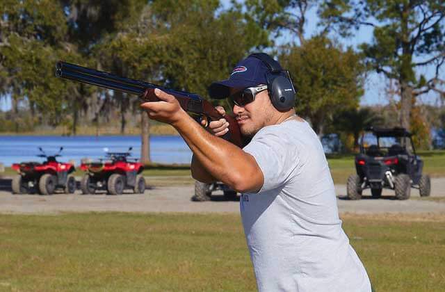man aims shotgun at clay pigeon with atvs and lake in the background at revolution adventures clermont