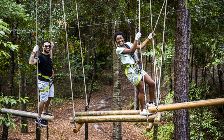 man and woman in climbing gear walking along suspended logs above ground with tree canopy at orlando tree trek