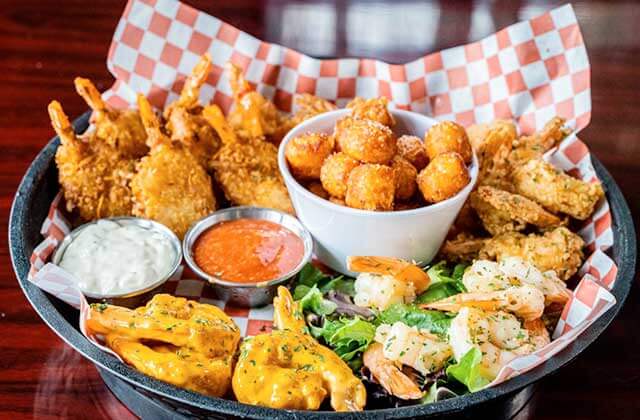 black woven basket with red and white checkered paper holding fried shrimp, fried okra, assorted cooked shrimp and seafood with tartar and cocktail sauces in bowls at Nantucket Shrimp Shack in Kissimmee, Florida.