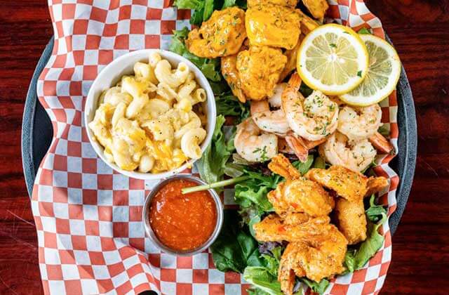 arial view of a black platter with a red and white checkered wax paper holding a bowl of macaroni and cheese, tartar sauce and three types of fried and steamed shrimps with lemon and lettuce garnish at Nantucket Shrimp Shack in Kissimmee, Florida.