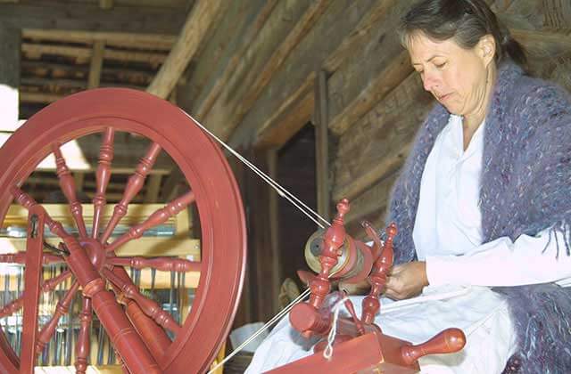 woman in a period costume using a spinning wheel on the porch of a log cabin at tallahassee museum florida