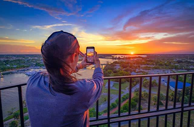 woman takes photo with phone of orange and blue sunset over inlet from lighthouse balcony at jupiter inlet lighthouse museum florida