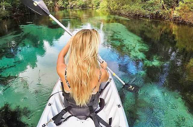 woman in kayak paddling through scenic springs-fed river looking for manatees with clear green water and dense tropical foliage for floridas adventure coast at weeki wachee destination feature