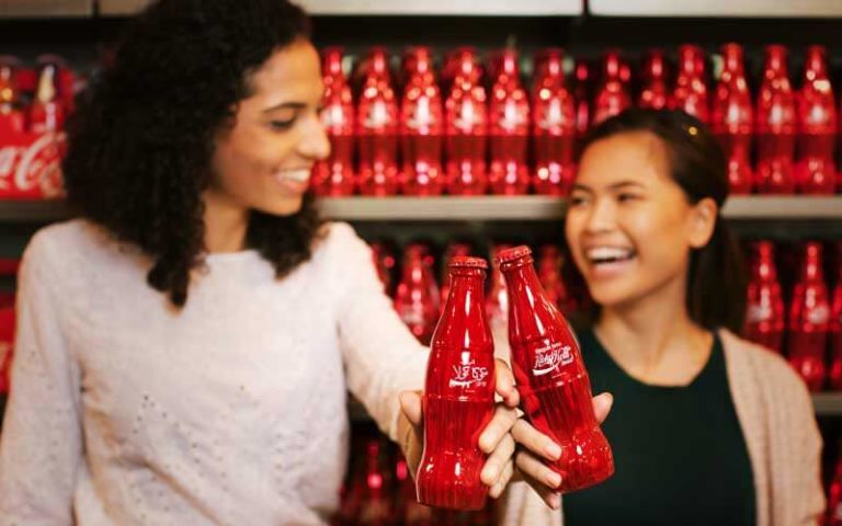 two girls toasting red metal coke bottles with retail shelf behind at coca cola store at disney springs