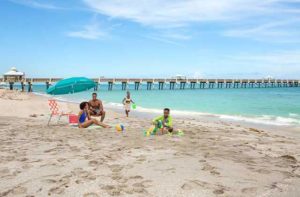 sunny beach with family of four under a blue umbrella with pier behind at the palm beaches florida