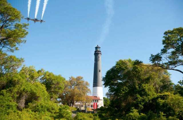 airplanes fly over a black and white lighthouse with trees at pensacola lighthouse florida