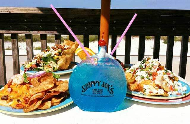blue fishbowl drink with pink straws with three seafood entrees on an outdoor table at sloppy joes treasure island