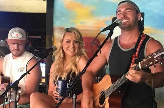 two men and a woman sing and play on stage with a colorful backdrop at sandbar sports grill key west