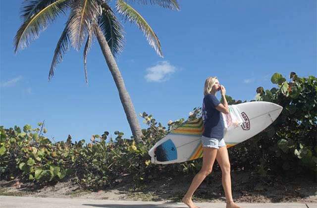 woman walks with surf board along beach with palm tree for ron jon surf shop panama city