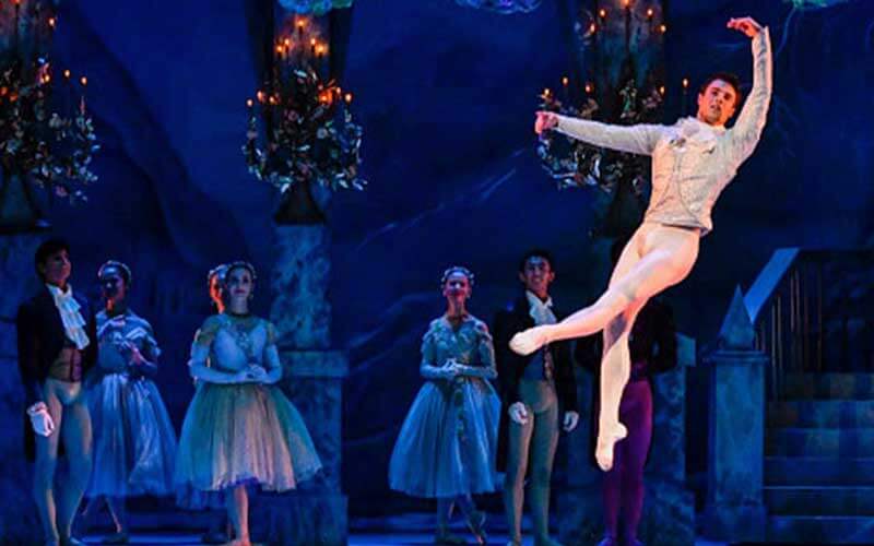 male dancer leaps on stage in front of female dancers wearing dresses 