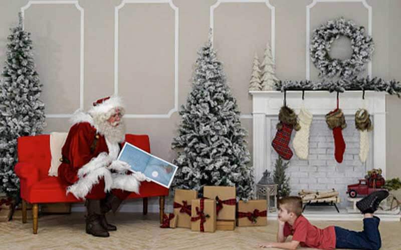 santa claus reads a story book to a boy in a christmas setting with trees fireplace stockings and gifts