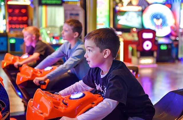 three boys compete in a motorcycle race game with many arcade games in the background at fun games arcade kissimmee