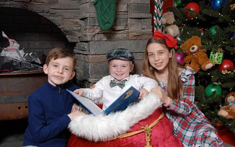 three children read a book in holiday attire in front of a brick fireplace