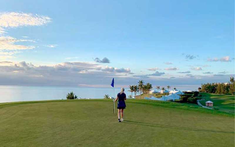 woman holds golf flag in the middle of a rolling golf course with puffy white clouds and clear blue skies