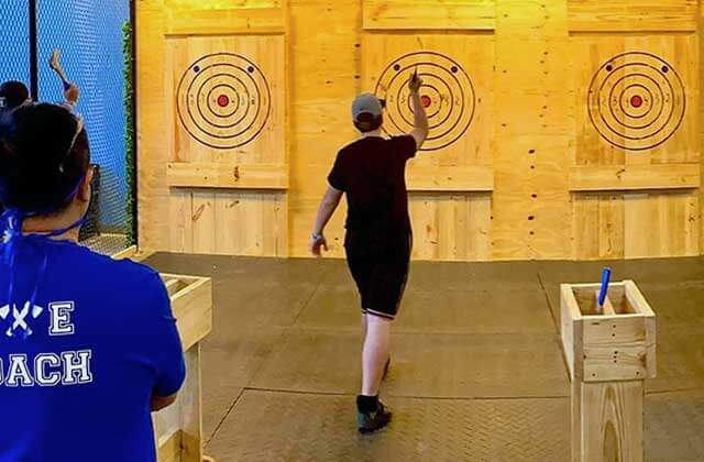 two men throwing axes at a wooden wall of targets with an axe coach watching at dezerland park orlando