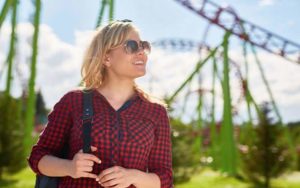 blonde woman with aviator sunglasses red checkered shirt and shoulder strap bag with roller coasters in background