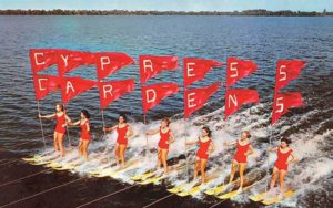 historic photo of seven woman water skiers in red bathing suits with red flags spelling cypress gardens on opening day