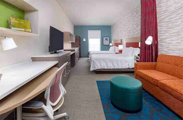hotel room with two large beds sofa desk and colorful accents at home2 suites by hilton orlando near universal