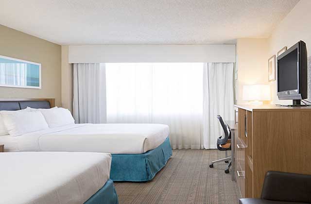 large hotel room with two king size beds a large window desk and tv in brown with blue accents at holiday inn palm beach airport conference center