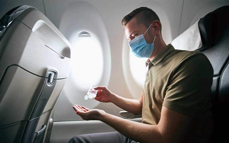 man in airplane seat wearing blue mask and applying sanitizer to hands