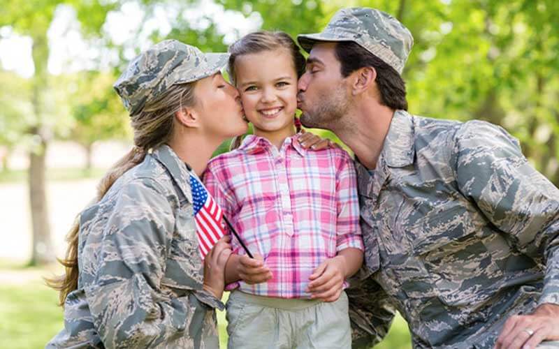 military mom and dad wearing uniforms and kissing little girl with a small american flag in her hand and trees in the background
