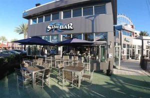 front exterior of gray building with patio seating dark blue umbrellas and observation wheel above right at i-drive starbar icon park orlando