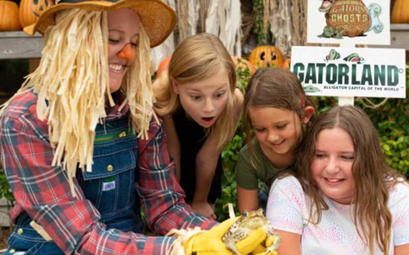 three young girls look in terror and joy at a frog held by a woman dressed as a scarecrow at the gators ghosts and goblins halloween fall and oktoberfest event at gatorland orlando