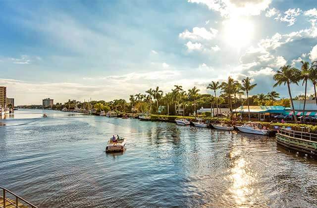 two people in a small yacht on inlet with docked boats along bank with palm trees and buildings at delray beach destination feature