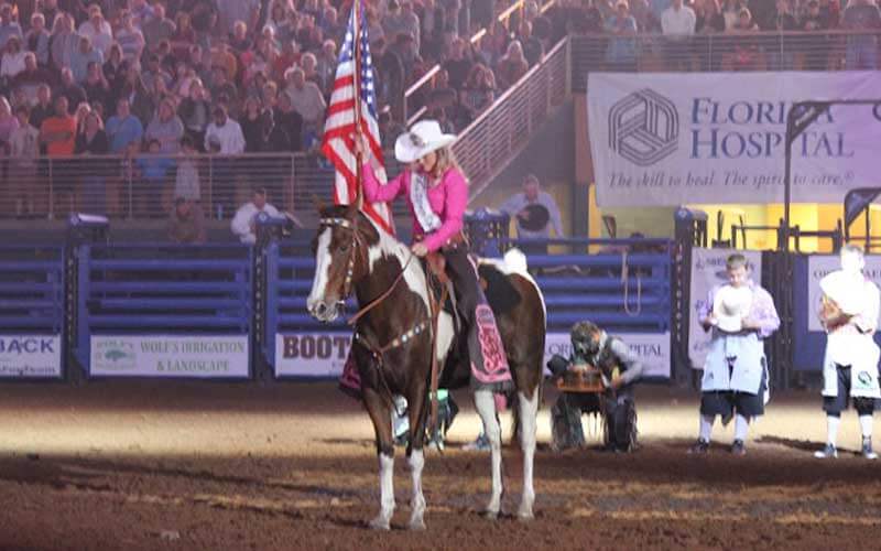 woman wearing pink cowboy apparel on horseback carrying flag in arena with crowds for silver spurs rodeo southern showdown blog