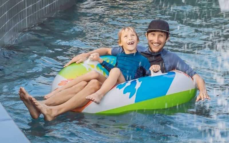 a small boy sits on a dads lap floating on an inner tube along a lazy river at a waterpark in florida 