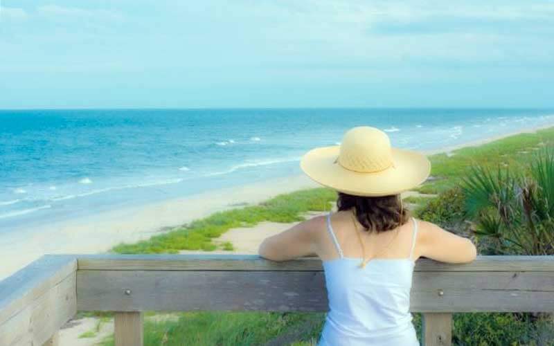 a woman wearing a tanktop and a straw hat leans on a wood railing while looking out over green shrubs next to tan sand and blue ocean 