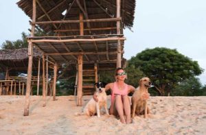 young woman wearing sunglasses and a pink shirt relaxes on a dog friendly beach in florida with a medium sized black and white dog and a large yellow dog in front of a tiki hut structure