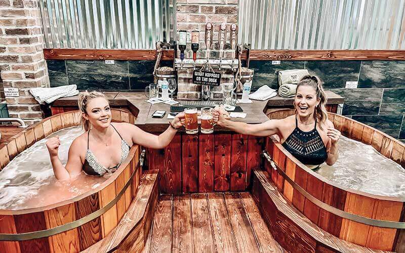 two girls wearing black bathing suits toast mugs of beer while sitting in large wooden beer barrel looking bathtubs with wood flooring wine and beer taps on a counter at beer spa orlando