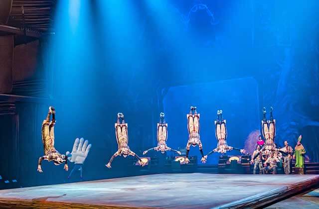 Acrobats mid flip with a person dressed as a hand in the background with people watching the acrobats in a blue foggy stage lighting at Cirque Du Soleil Disney Drawn To Life Show at Disney Springs Orlando.