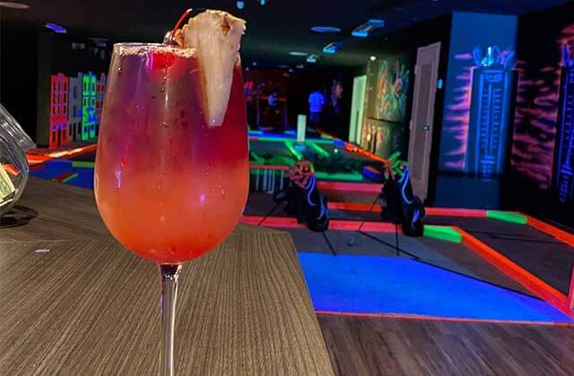 red cocktail with a cherry and a pineapple on the rim on a bar in front of a day glow neon colored putt putt golf course with neon colored art and graffiti on the walls and two golf bags full of glow in the dark putt putt golf clubs at dezerland park miami indoor mini golf attraction