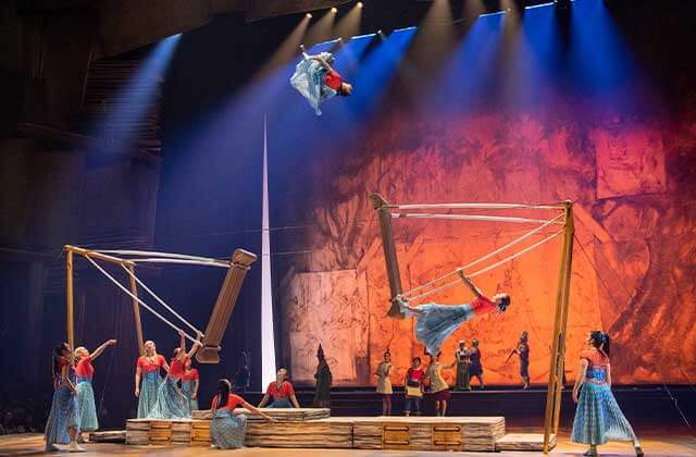 female acrobats wearing red and blue dresses on stage one is swinging and one is mid flip high in the air on stage at Cirque Du Soleil Disney Drawn To Life Show at Disney Springs Orlando.