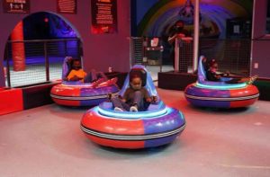 three children sit in red and purple bumper karts spinning on a bumper rink with red lighting at dezerland park miami indoor attraction