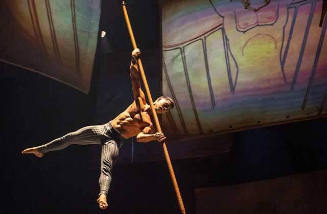 A shirtless man wearing brown striped pants holds himself up on a brown pole with a background of a mural of a sketched male torso at Cirque Du Soleil Disney Drawn To Life Show at Disney Springs Orlando