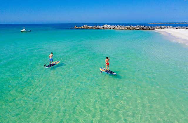 three people paddleboard on crystal clear blue water along a rock jetty at the st andrews shell island panama city beach florida