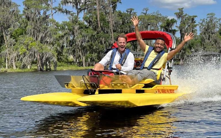 two men speeding across water in catboat with trees behind at catboat escapes