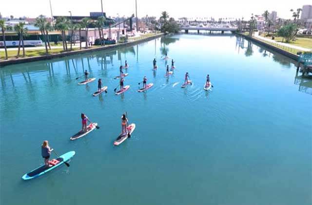 many people stand up on paddle boards in a canal surrounded by grass, palm trees, buildings on a stand up paddle board tour with three brothers adventures