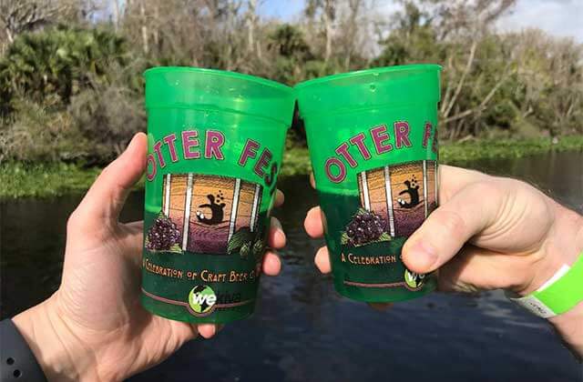 two hands toast beer in green otter fest cups in front of wekiva springs with trees in the background