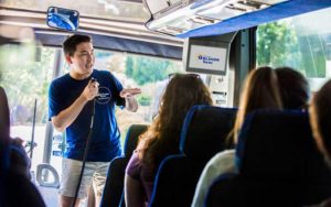 man stands speaking into a microphone at the front of a bus giving a tour of orlando with a television screen displaying the original orlando tours logo