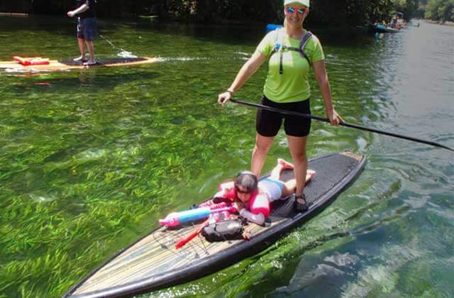 woman stands on stand up paddleboard over a child laying on the paddleboard with bags and items while floating down a river surrounded by green leaves and foliage next to a person on a stand up paddleboard