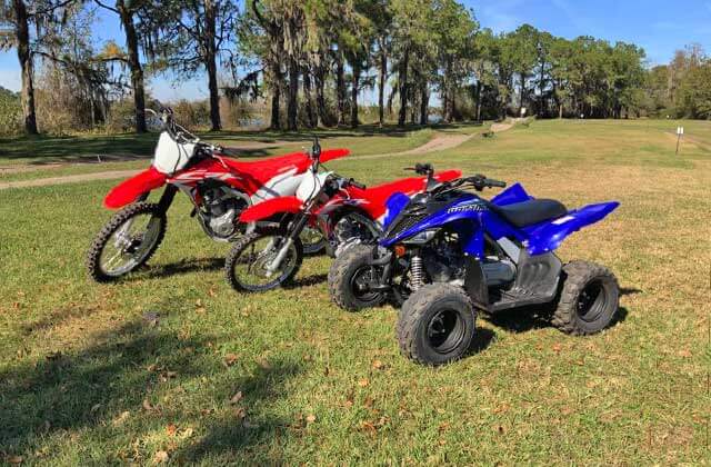 two red youth size dirtbikes and a blue youth size atv parked on a large grassy lot with trees and lake at revolution rentals at bone valley atv park