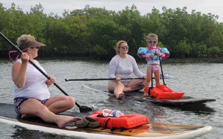 two ladies and toddler on paddleboards on water with trees behind at three brothers boards