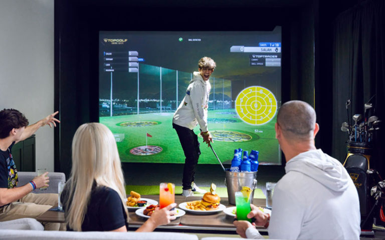 family cheering while boy swings club and table eating large screen game at topgolf swing suites