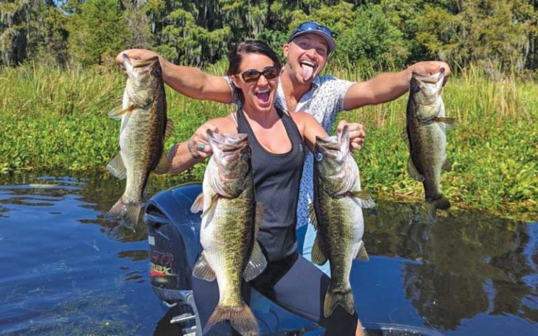 man and woman standing on boat with trees showing off four big bass fish at david paycheck fishing