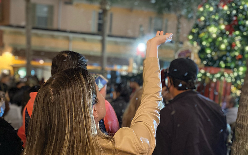 woman in crowd holding hand up to catch snow with street and christmas tree at night for now snowing event at celebration town center