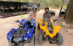 father and son on atvs with helmets and black trailer behind and trees at moore atv rentals